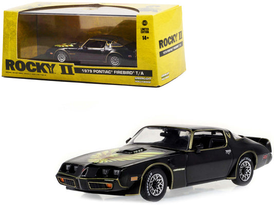 A diecast model of a 1979 Pontiac Firebird Trans Am in black color with hood, positioned in front of its yellow box packaging labeled 'Rocky II'.