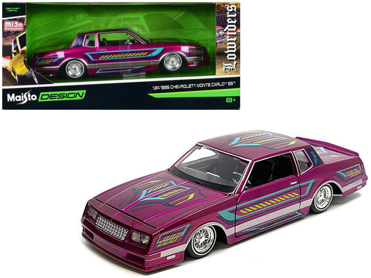A diecast model of a 1986 Chevrolet Monte Carlo SS Lowrider in pink with graphics, featuring real rubber tires and detailed compartments.