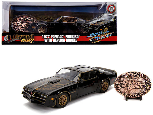 A 1:24 scale die cast model of a 1977 Pontiac Firebird Trans Am in black color, accompanied by a replica buckle from the movie 'Smokey and the Bandit'. The model has real rubber tires and is displayed in its original packaging.