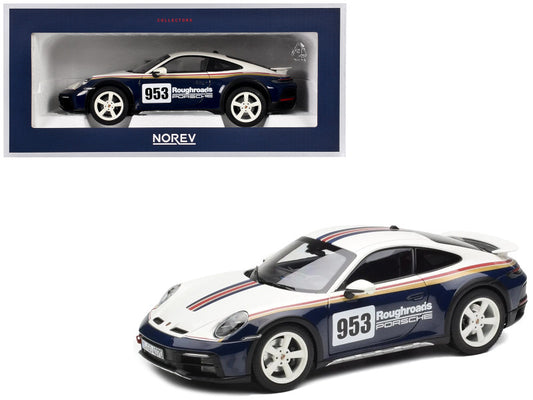 A 1/18 scale diecast model of a blue and white 2023 Porsche 911 Dakar with the number 953, displayed in its original packaging.